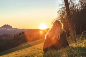Woman sitting on a grassy hillside facing sunlight, accompanying a guide to hangxiety meaning.